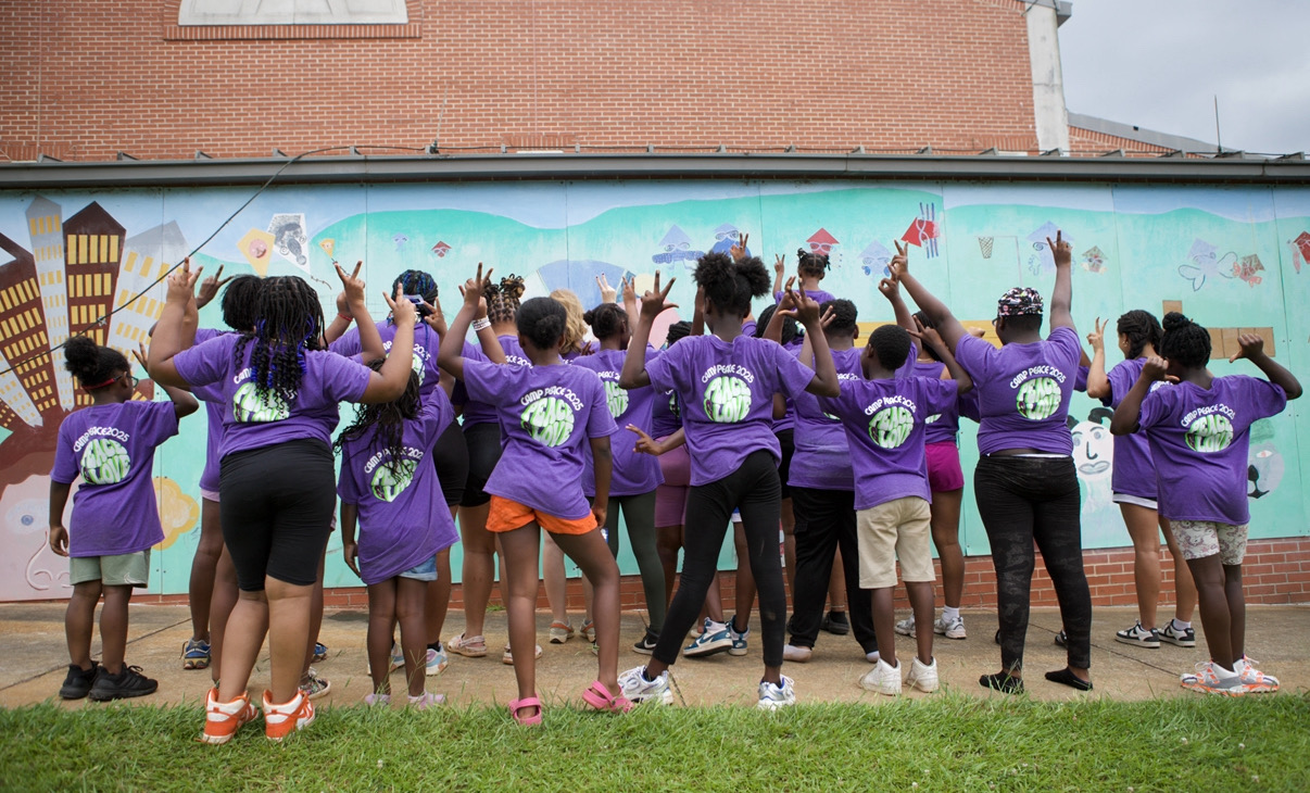 Campers in purple Camp PEACE shirts cheering together in front of a colorful mural
