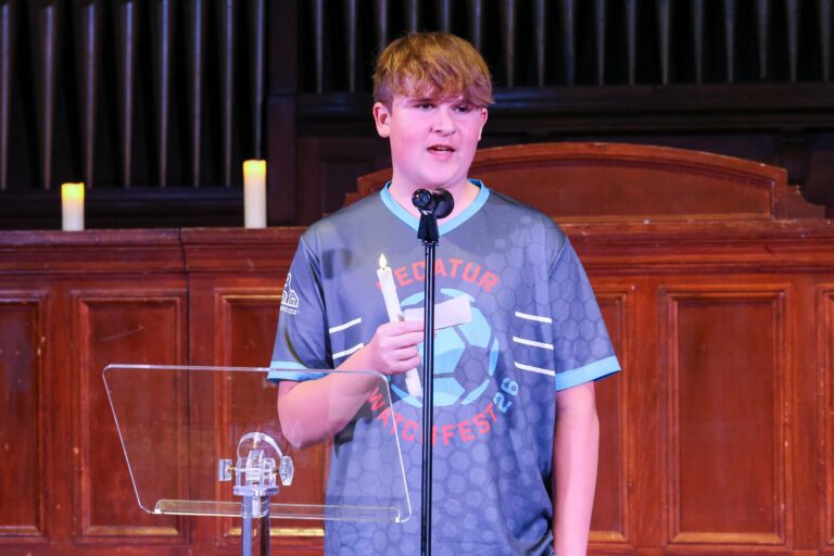 A young speaker shares remarks at the podium, holding a candle