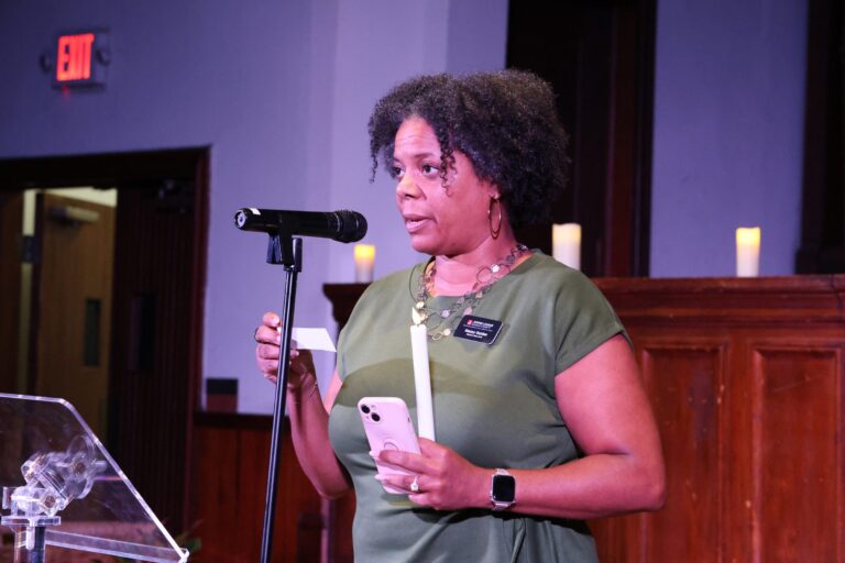 A speaker reads remarks at the microphone, holding a candle